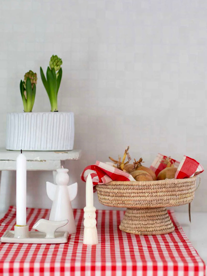 Decorative items on a table with a red and white checkered tablecloth, including a white cake stand with plants, a basket with bread, and a small angel sculpture.