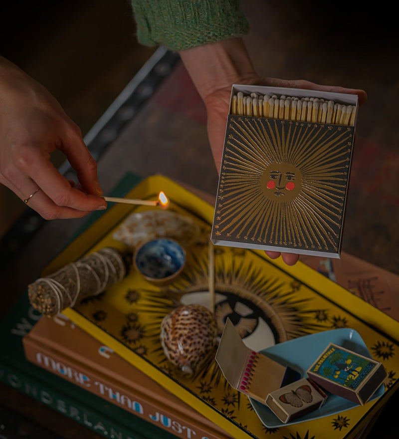 Person lighting a match from a decorative matchbox with a sun design on a table with books and other items.
