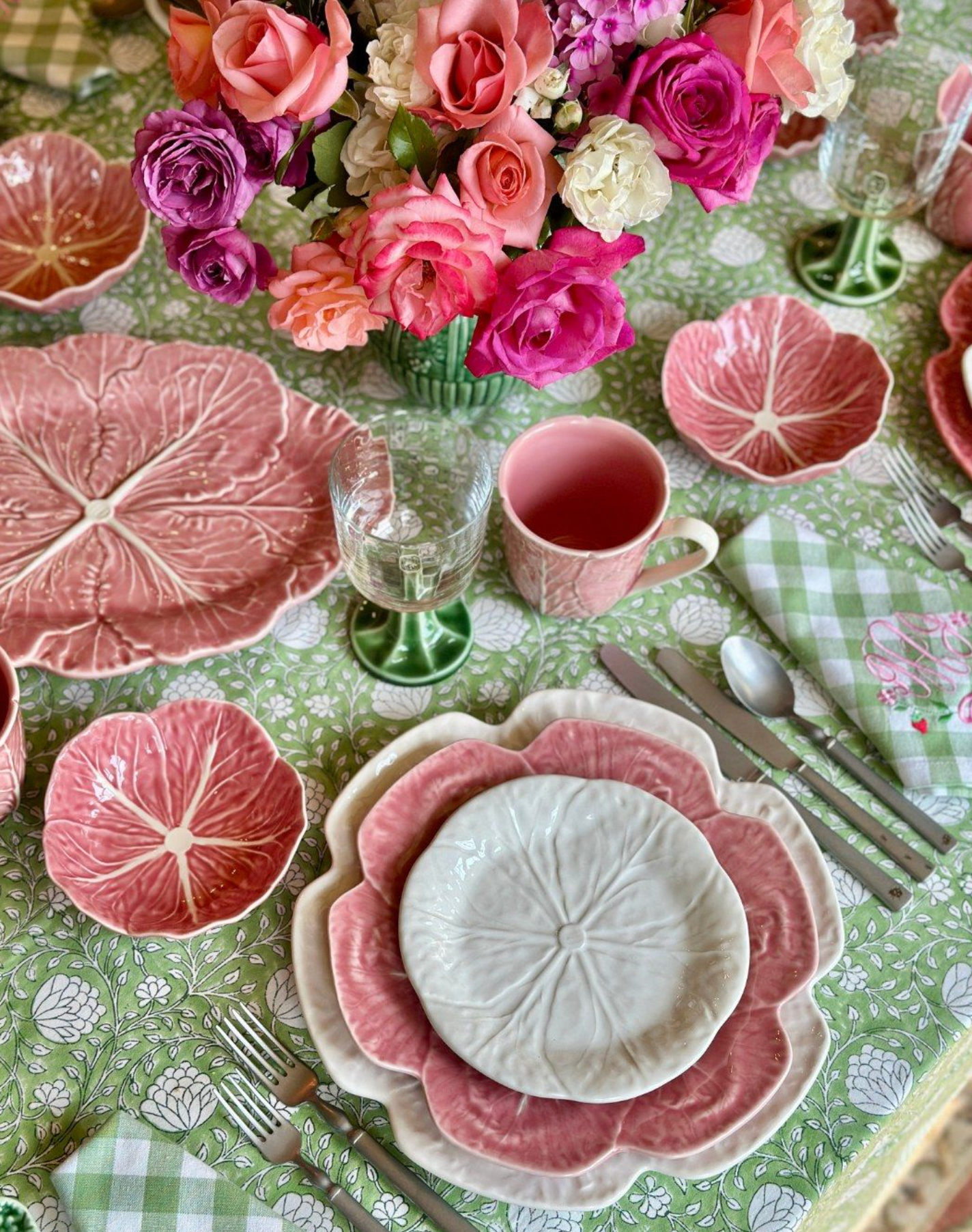 Decorative table setting with pink cabbage plates, cups, and a vase of flowers on a green patterned tablecloth.