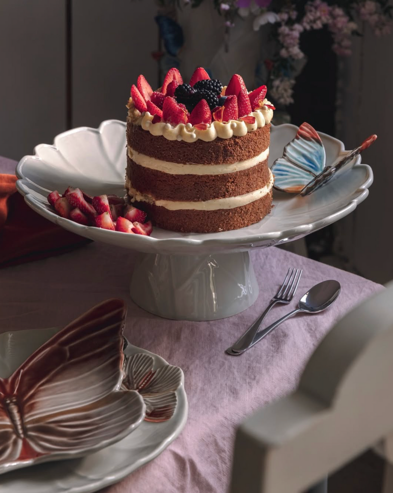 Cake with strawberries and cream on a white cake stand on a table with butterfly-themed plates.