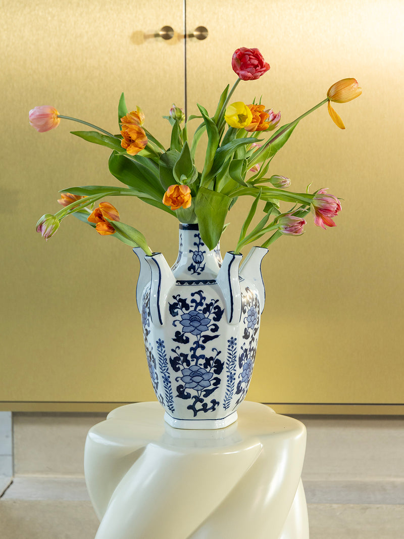 A white ceramic vase with blue floral patterns, filled with tulips in pink, orange, yellow, and red, displayed on a white sculptural pedestal against a warm beige wall.