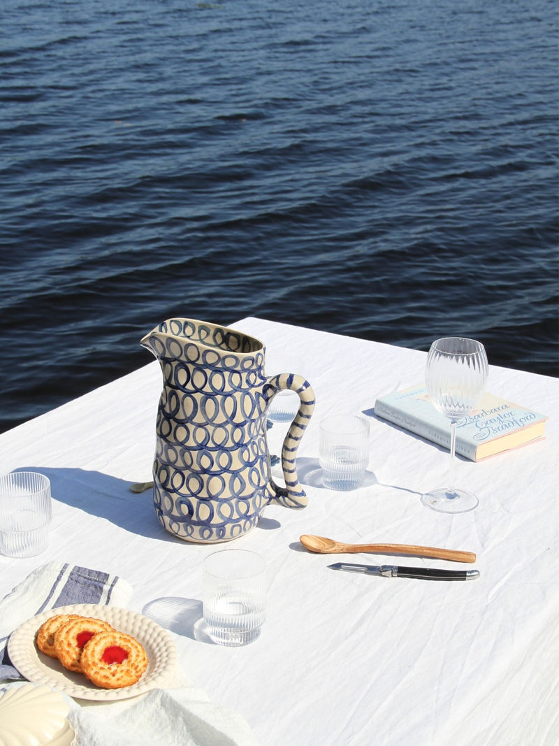 A patterned ceramic pitcher with blue and cream circles sits on a white tablecloth next to an ocean view, with wine glasses, a wooden spoon, and cookies nearby.
