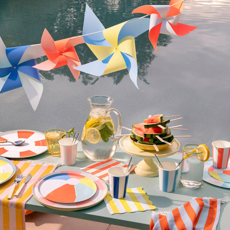Outdoor table setting with colorful plates, cups, and watermelon skewers, featuring a pool in the background.