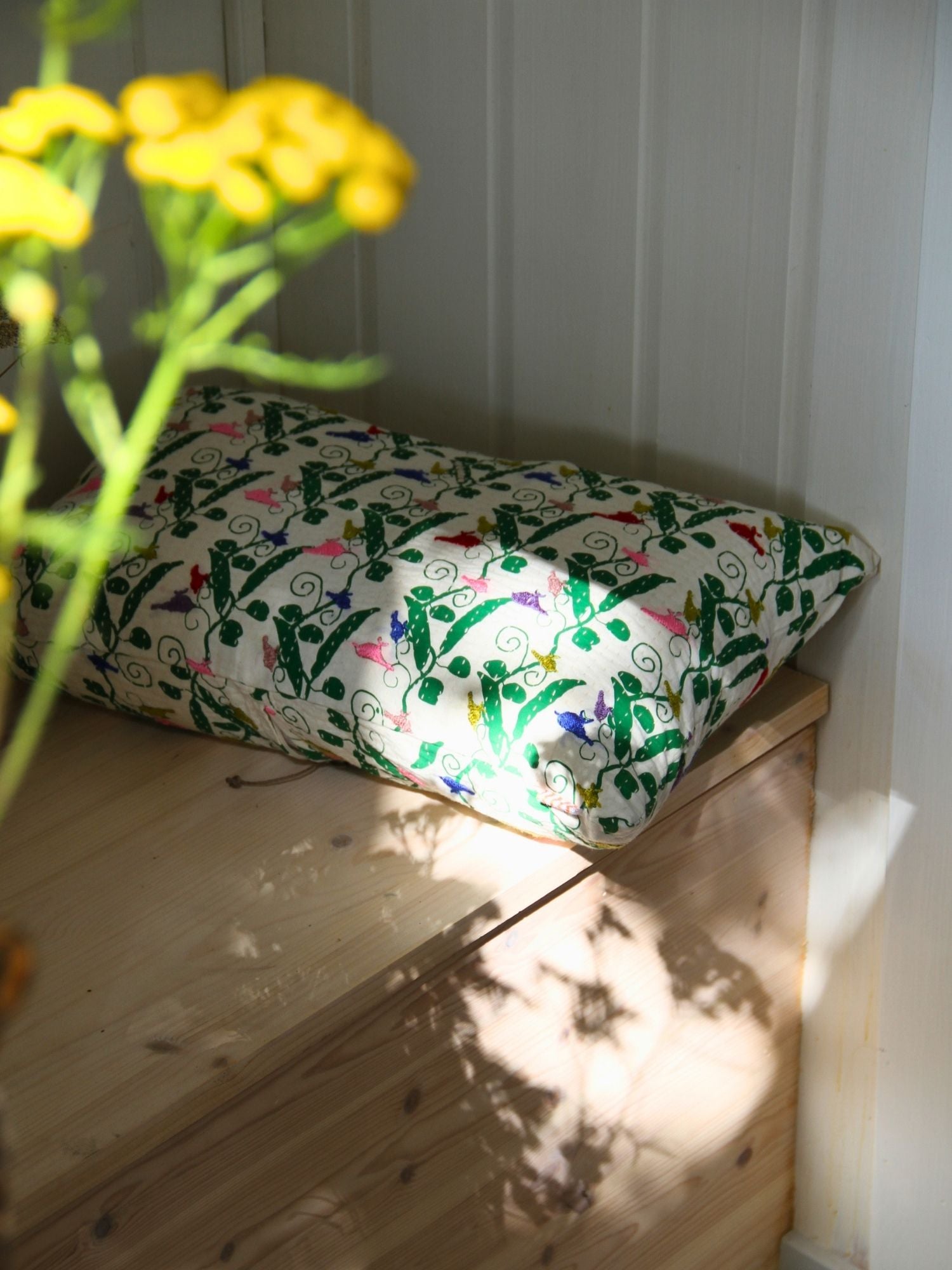 A rectangular decorative pillow with a colorful floral and vine pattern on a cream background, resting on a light wood bench in natural sunlight with yellow flowers in the foreground.