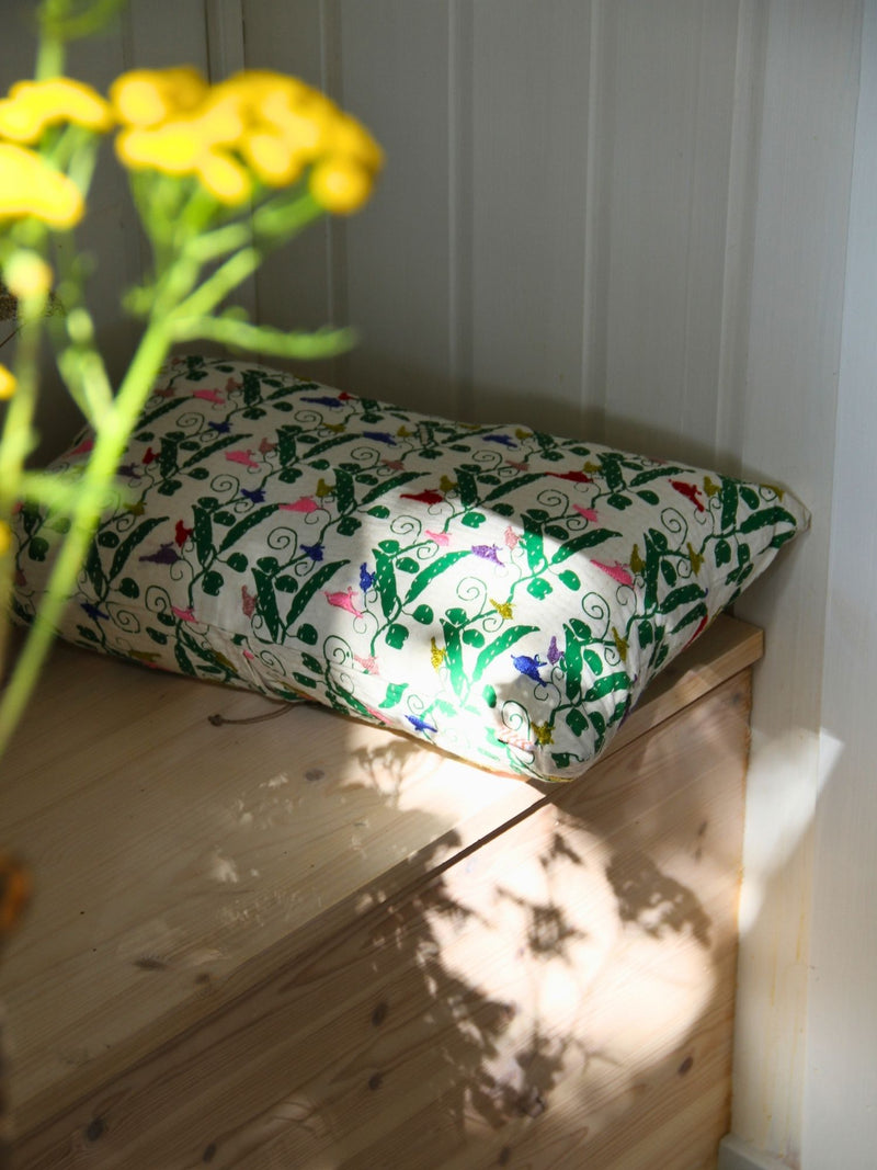 A rectangular decorative pillow with a colorful floral and vine pattern on a cream background, resting on a light wood bench in natural sunlight with yellow flowers in the foreground.