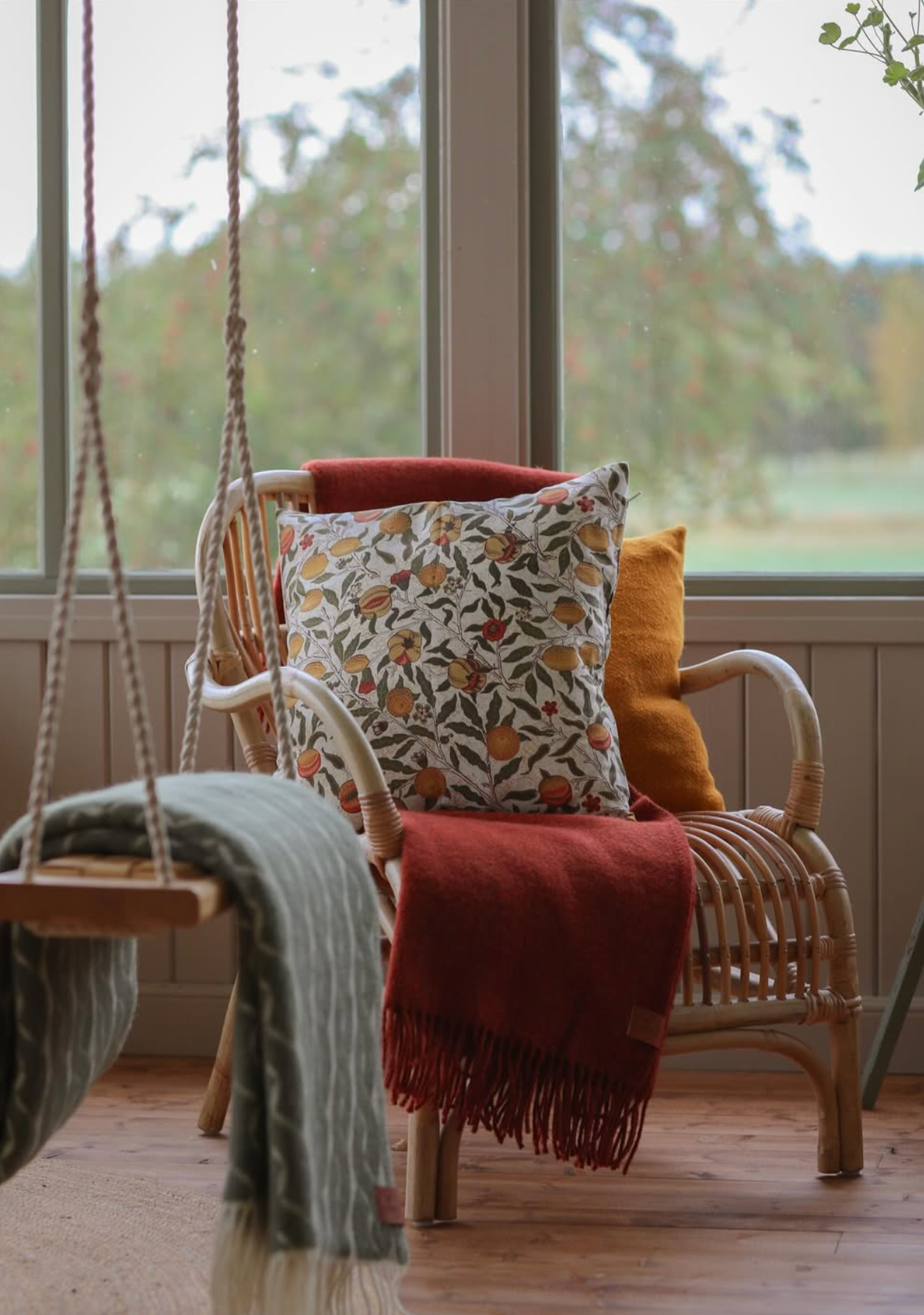 Rattan chair with William Morris print patterned cushion and a blanket in a room with large windows.