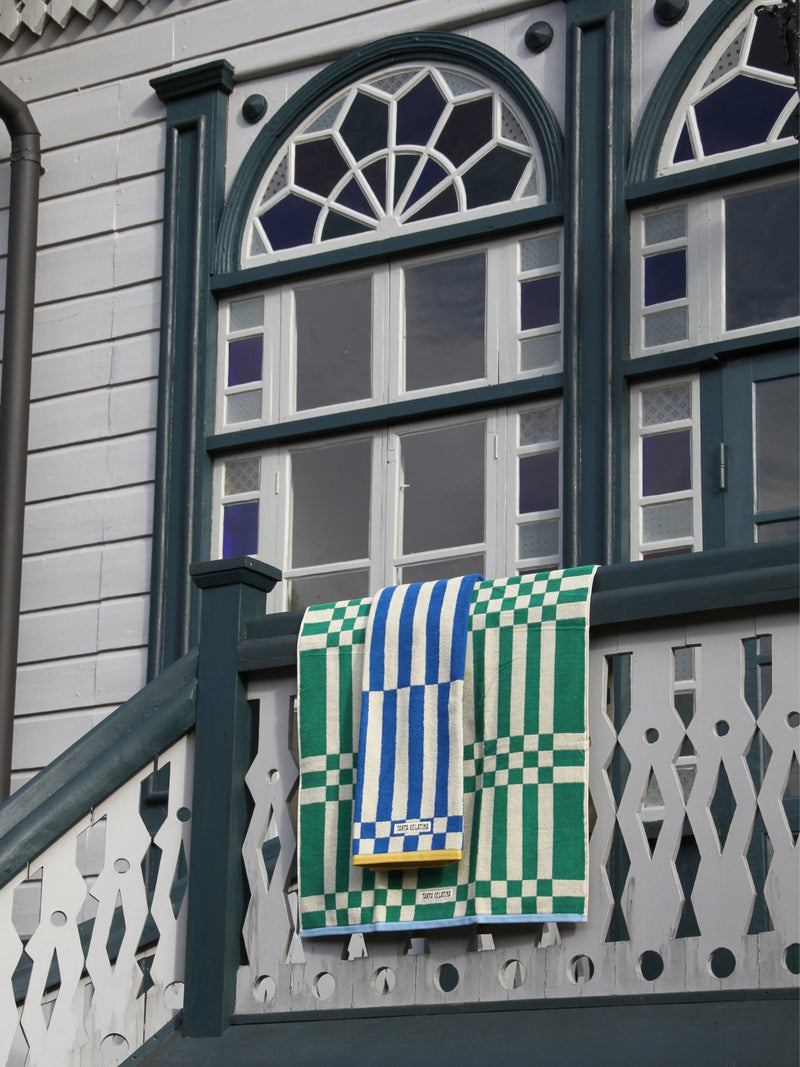 A green and white checkered towel with blue and white striped patches hanging from a dark green wooden balcony railing of a house with white siding and decorative windows.
