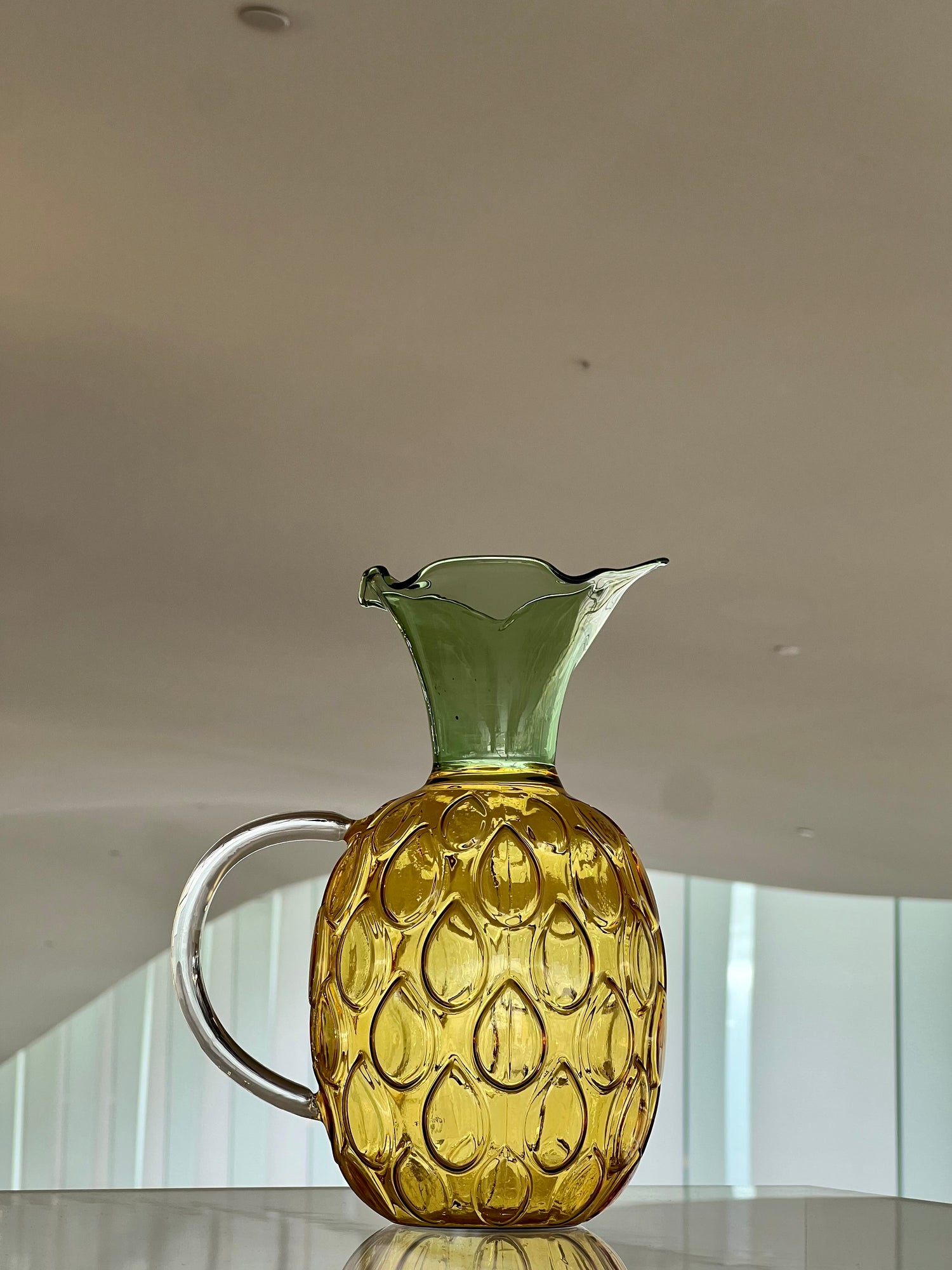 Glass pitcher shaped like a pineapple on a reflective surface with a neutral background