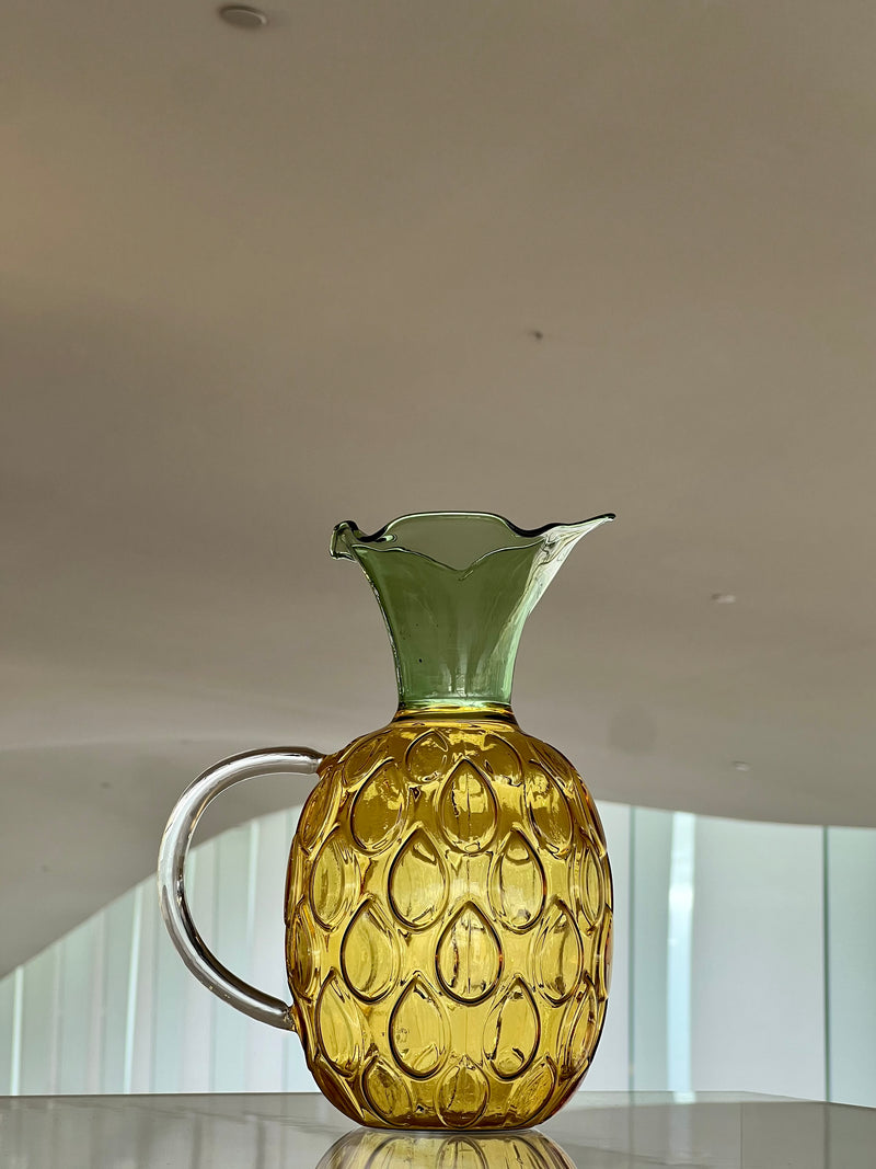 Glass pitcher shaped like a pineapple on a reflective surface with a neutral background
