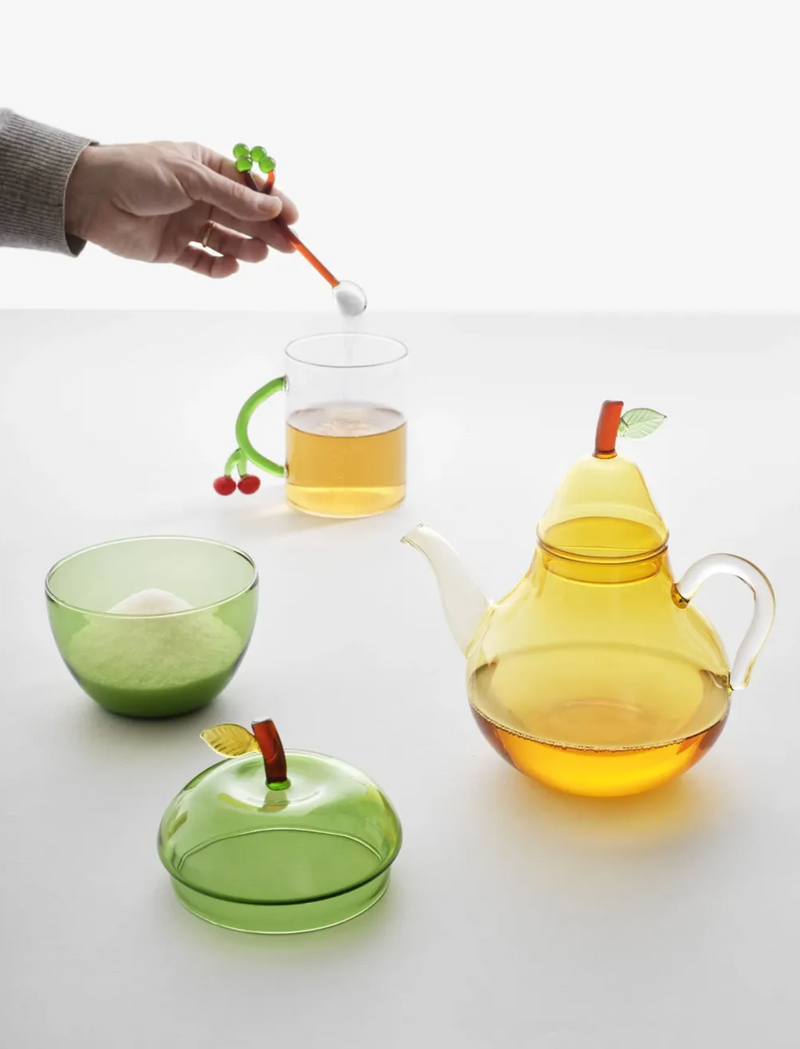 A glass teapot and matching cup set in translucent yellow-green tones with apple-shaped lids and stems, shown on a white surface. A hand is visible holding a spoon above a clear glass cup containing tea.