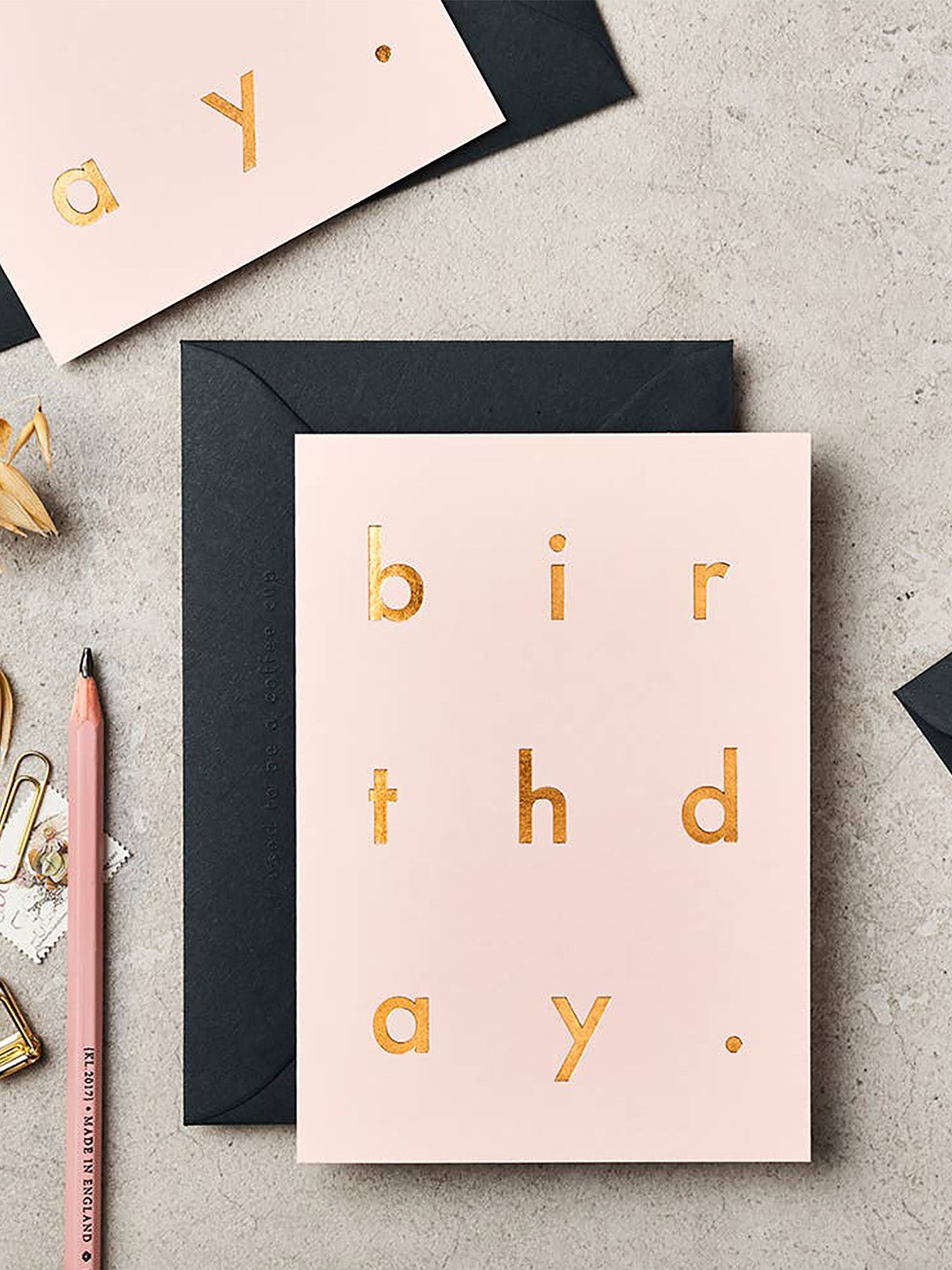 A flat-lay of a light pink birthday card with gold foil letters spelling 'birthday.' placed on top of a black envelope, with additional matching stationery and a pencil on a gray textured surface.