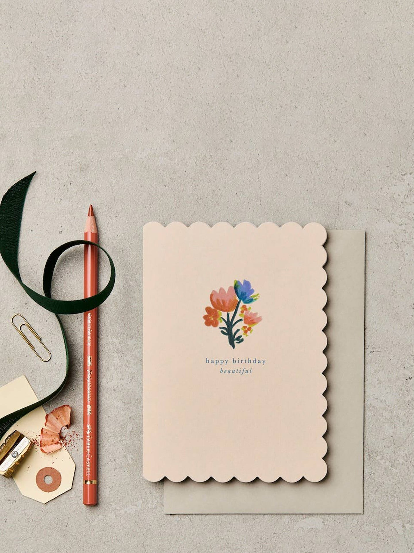 A scalloped-edge birthday card with a colorful floral illustration and the words 'happy birthday beautiful,' shown with a matching envelope on a light gray textured surface alongside a soft grey envelope pencil, ribbon, paperclip, and small decorative items.