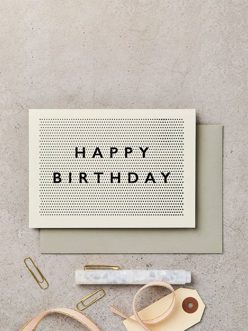 A square cream birthday card with the words 'HAPPY BIRTHDAY' in black serif letters over a dotted pattern, placed on top of a muted sage envelope. The card is styled on a light gray textured surface with gold paperclips, a marble pen, ribbon, and a gift tag.