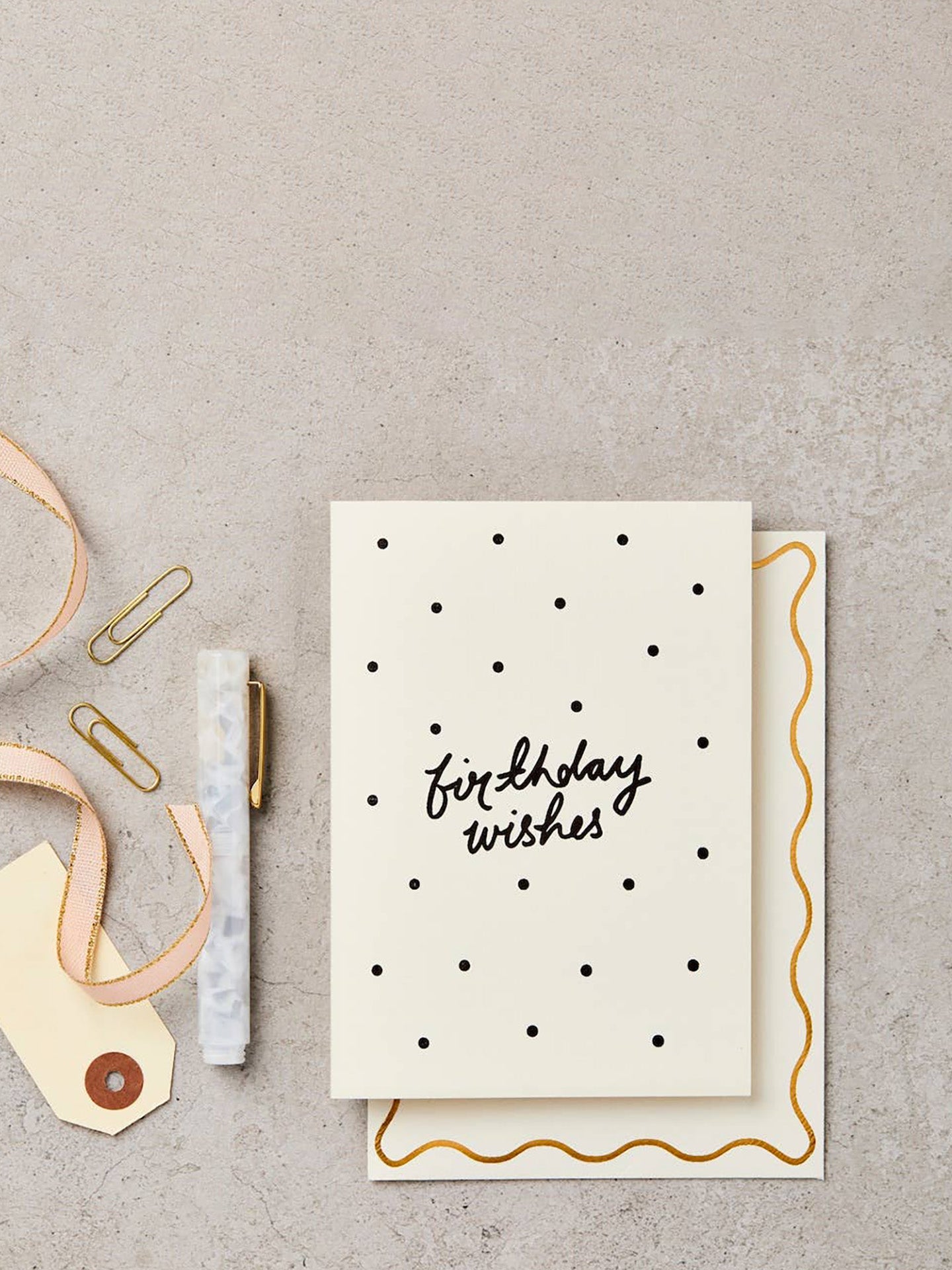 A cream-colored birthday card with black handwritten text reading 'birthday wishes' and black polka dots, shown with a matching envelope on a textured gray surface. A pen, paper clips, and a ribbon are arranged around it as styled flat-lay props.