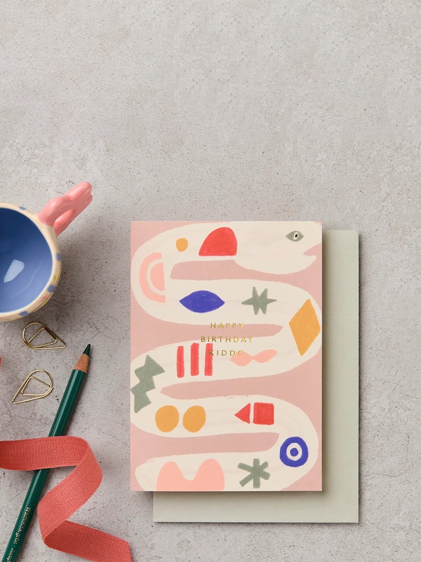 A light pink greeting card with a curvy snake illustration and gold 'Happy birthday kiddo' text, placed on a light gray surface with a matching envelope, a blue mug, a green pencil, and a coral ribbon nearby.