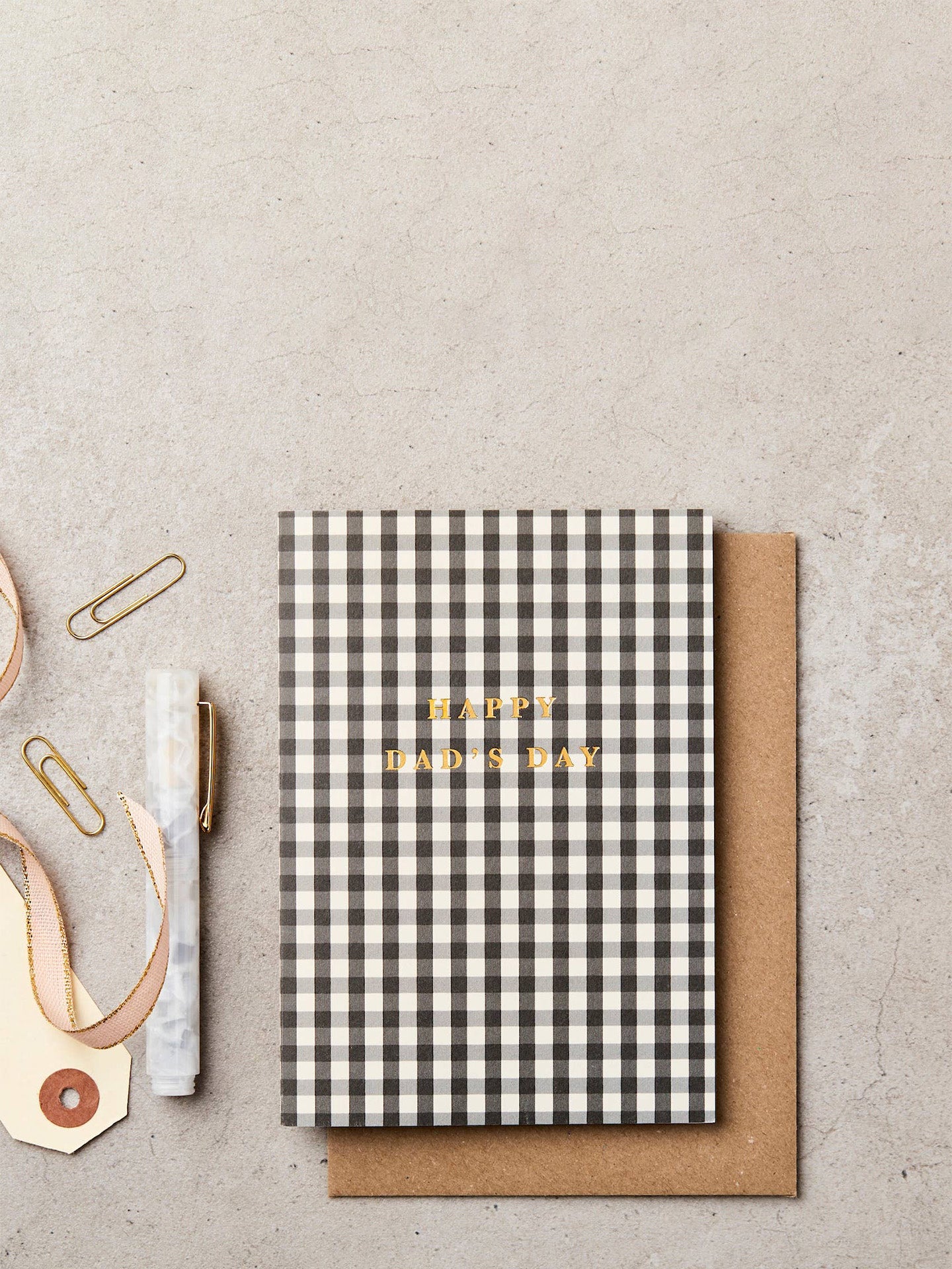 A black and white gingham check notebook with gold foil 'HAPPY DAD'S DAY' text, displayed with a brown kraft envelope with gold paperclips, a marble pen, and a ribbon on a textured beige background.