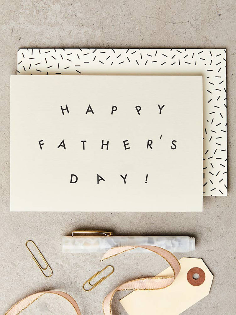 A Father's Day greeting card with the words 'HAPPY FATHER'S DAY!' in black lettering on an off-white background, layered over a patterned envelope. The card is styled on a textured gray surface with gold paperclips, a marble pen, and a ribbon tag.