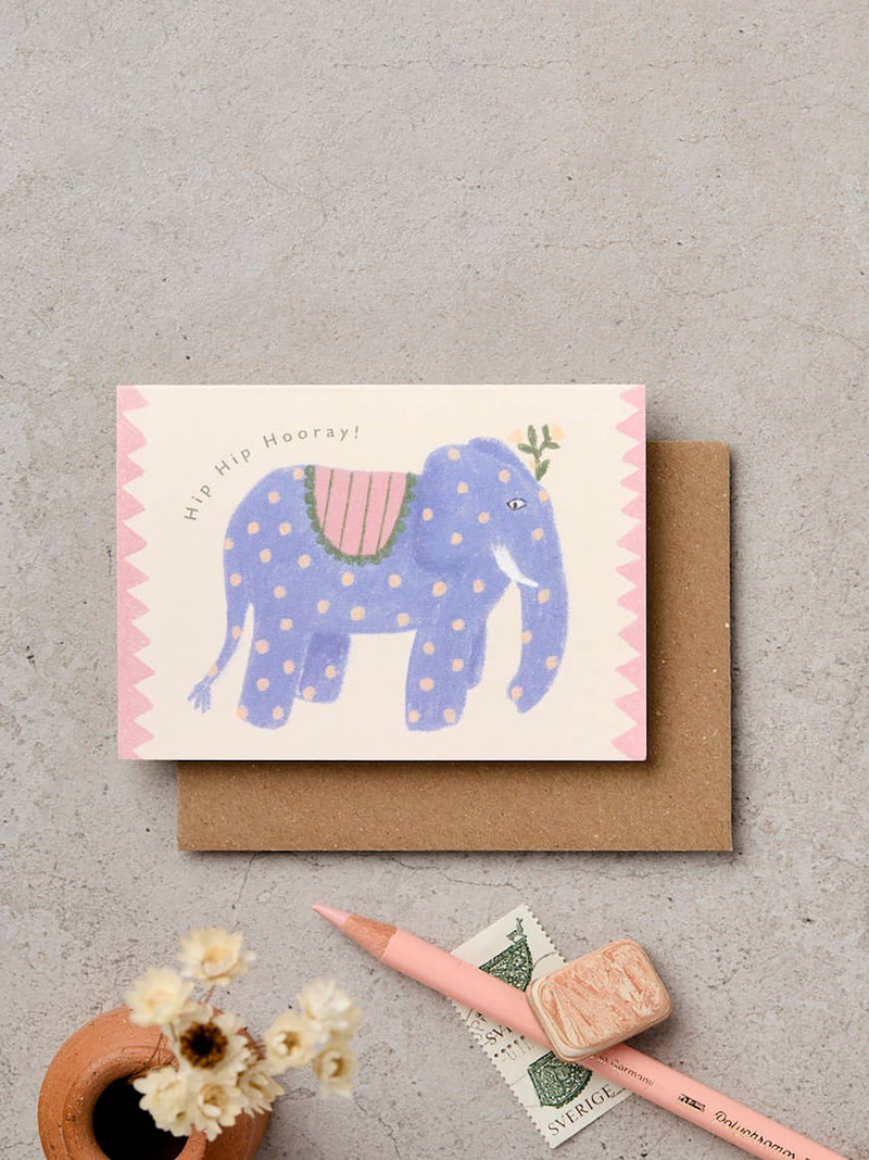 A greeting card featuring a whimsical blue elephant with pink polka dots and a pink saddle, placed on top of a brown envelope on a textured gray surface. A pink pencil, a small ceramic pot with dried flowers, and a postage stamp are arranged around it.