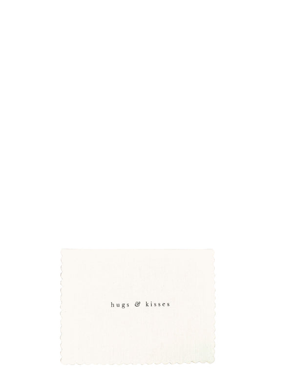 A square cream-colored fabric napkin with scalloped edges and the text 'hugs & kisses' printed in a small black serif font, set against a solid white background.