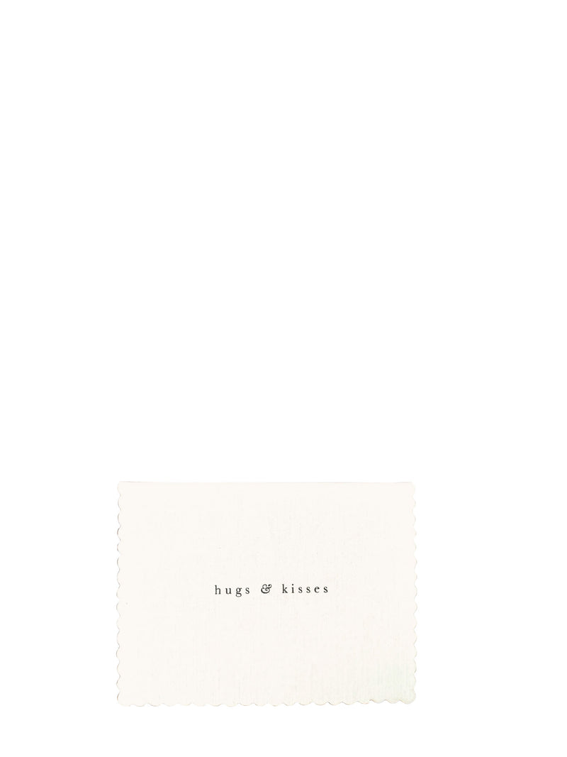 A square cream-colored fabric napkin with scalloped edges and the text 'hugs & kisses' printed in a small black serif font, set against a solid white background.