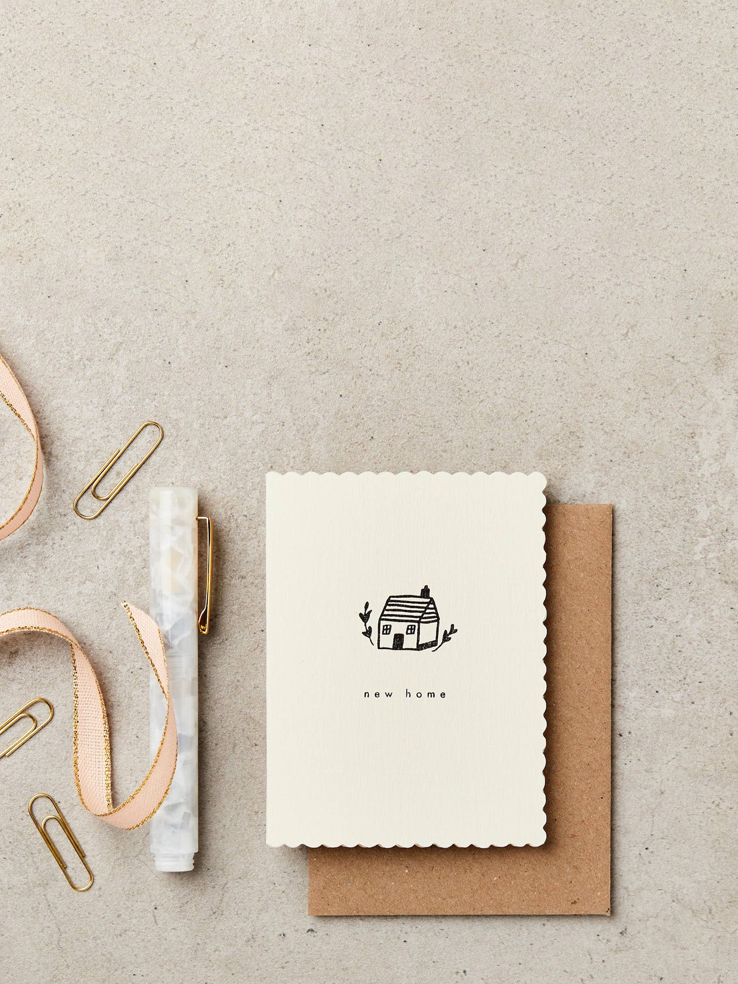 A cute off-white minimalistic greeting card with scalloped edges illustrated with an ink line drawing of a small cottage and text new home printed on black laying on concrete surface on top of a kraft brown envelope, next to a pen, some paper clips and a pink gift ribbon.