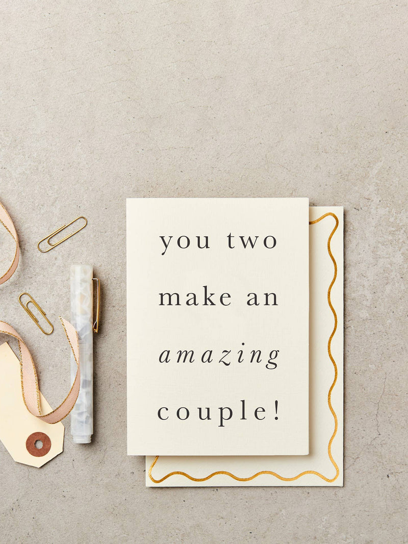 A square cream greeting card with black serif text reading 'you two make an amazing couple!' placed on top of a matching envelope with a gold wavy border, styled on a light gray textured surface with paper clips, a ribbon tag, and a marble pen.