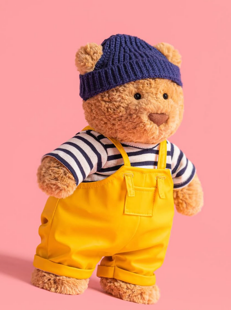 Teddy bear wearing a blue knitted hat, striped shirt, and yellow overalls on a pink background