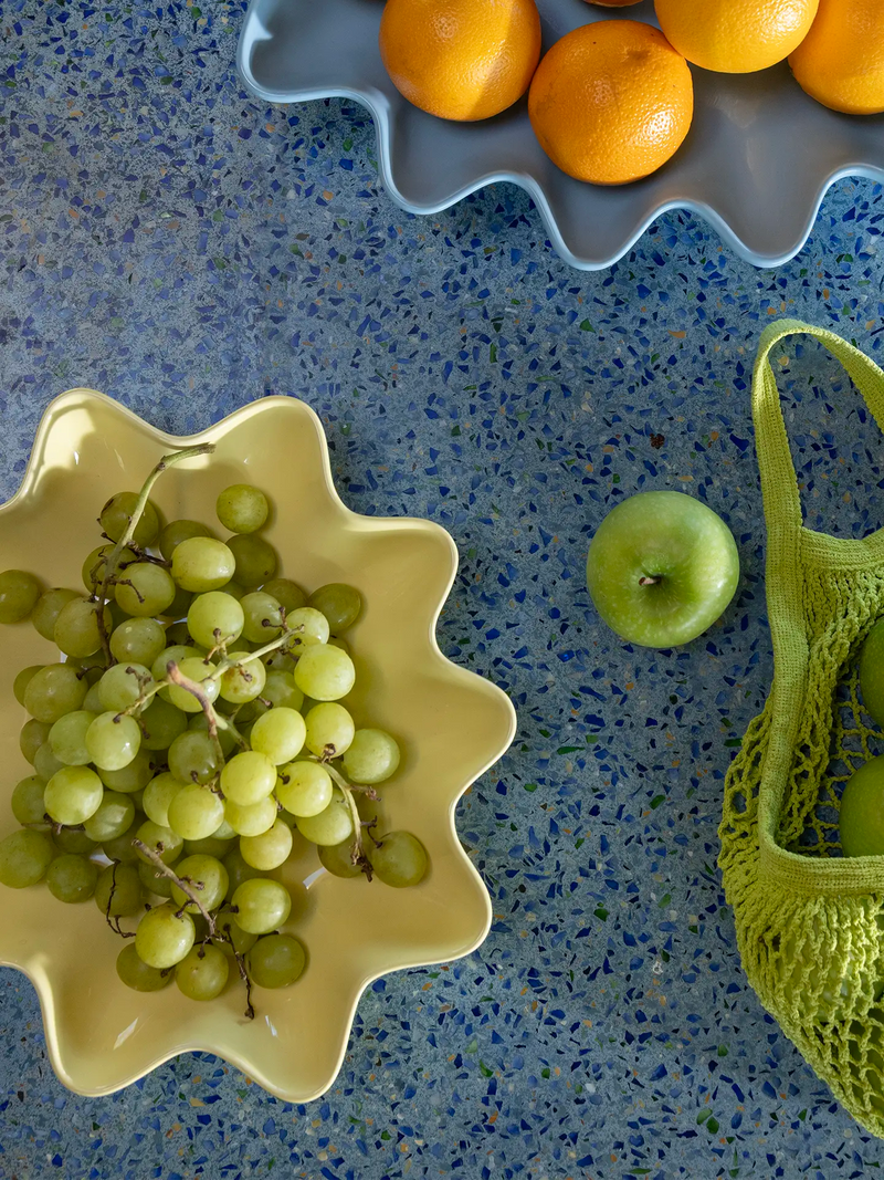 A styled overhead flat-lay of fresh fruit on a speckled blue countertop, featuring a yellow oval-shaped scalloped bowl filled with green grapes, a green apple, oranges in a blue scalloped dish, and a green mesh bag.