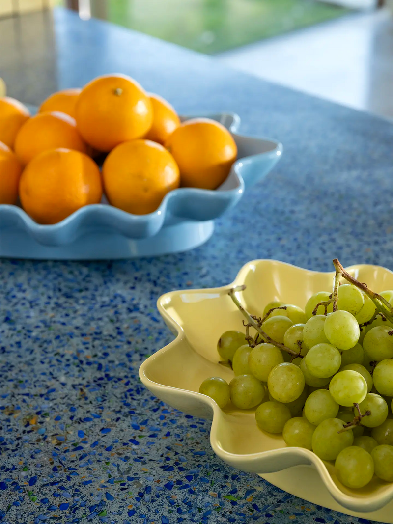 A yellow ceramic star-shaped bowl filled with green grapes on a speckled blue countertop, with a blue scalloped bowl of oranges blurred in the background.