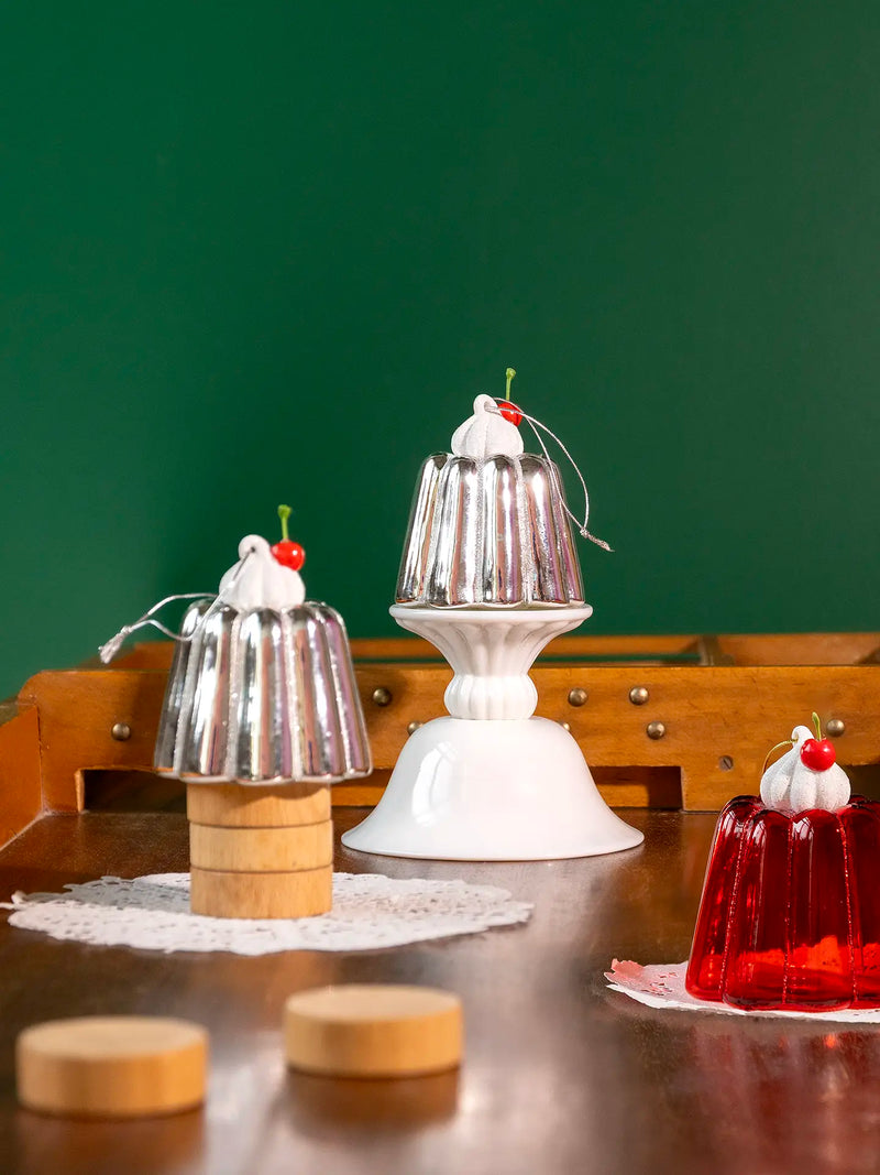 Three dessert-themed decorative objects displayed on a wooden table against a dark green background: a small silver gelatin mold on a wooden base with whipped cream and a cherry, a larger silver gelatin mold on a white pedestal stand with whipped cream and a cherry, and a red translucent gelatin mold with whipped cream and a cherry.