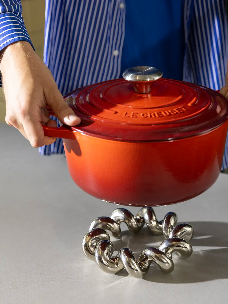 A person in a blue striped shirt holds a red Le Creuset enameled cast iron pot with its lid on, positioned above a chunky silver metal trivet on a light gray surface.