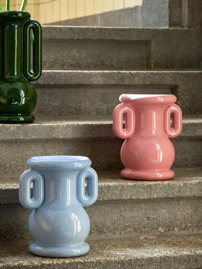 Two glossy ceramic urn-shaped planters with side handles, one in light blue and one in bubblegum pink, displayed on outdoor stone steps next to a dark green planter.
