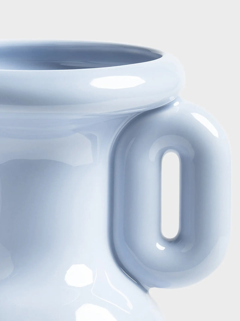 Close-up of a glossy ceramic pitcher or vase in a pale icy blue color, showing the thick integrated handle and smooth curved body against a light neutral background.
