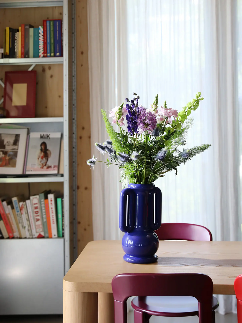 A vibrant blue ceramic vase filled with a colorful bouquet of purple, pink, and green flowers, set on a light wood dining table in a bright room with white curtains and a bookshelf in the background.