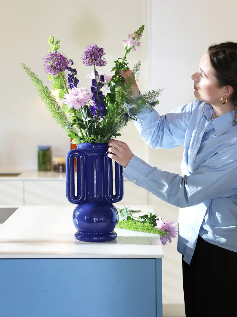 A woman in a light blue shirt and black pants adjusts a bouquet of purple, pink, and green flowers in a glossy cobalt blue ribbed vase on a kitchen counter.