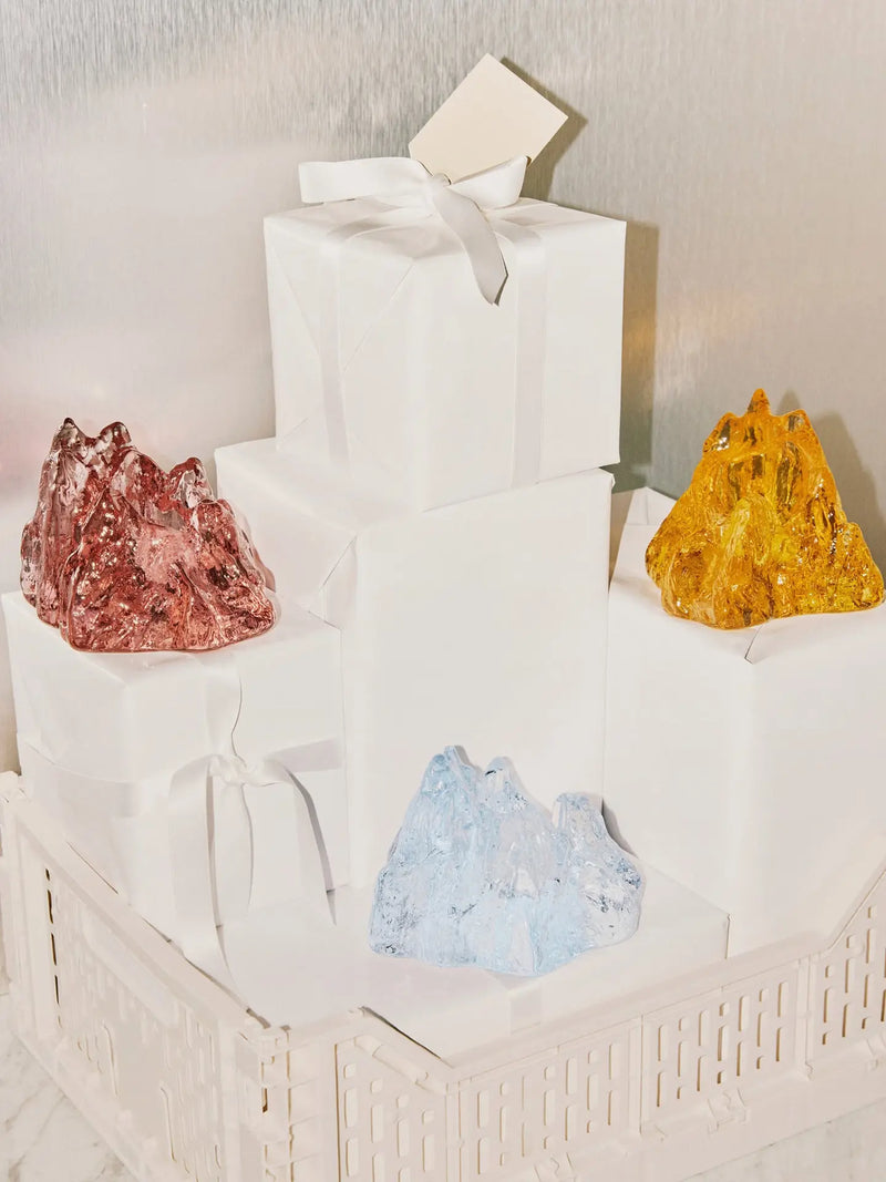 Four wrapped gift boxes stacked in a pyramid arrangement, with four raw crystal formations of tealight holders in rose pink, amber yellow, and light blue,  placed on and around the boxes.