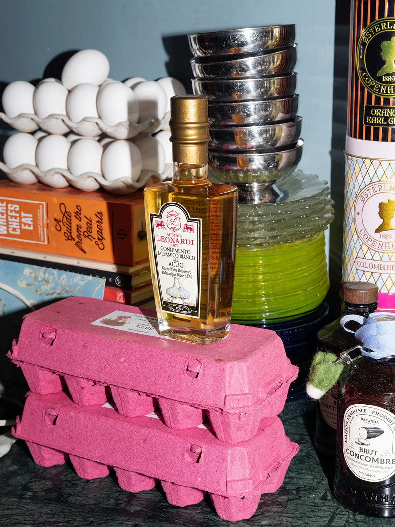 A bottle of Leonardi's GArlic infused white balsamic vinegar centered on a stack of pink egg cartons, surrounded by kitchen items including a carton of eggs, metal dessert bowls, a green glass plate stack, and other bottles in a dark green marble kitchen counter.