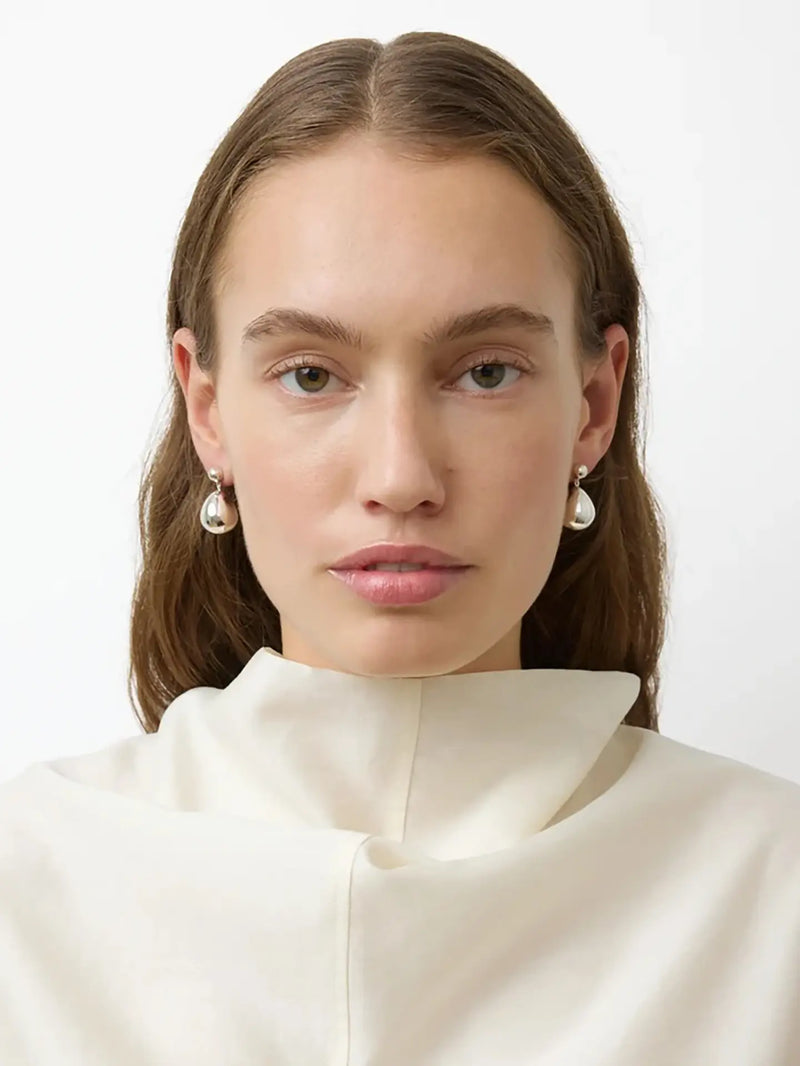 A close-up portrait of a woman with brown hair wearing small pearl-like silver drop earrings and a cream high-neck top against a plain light background.