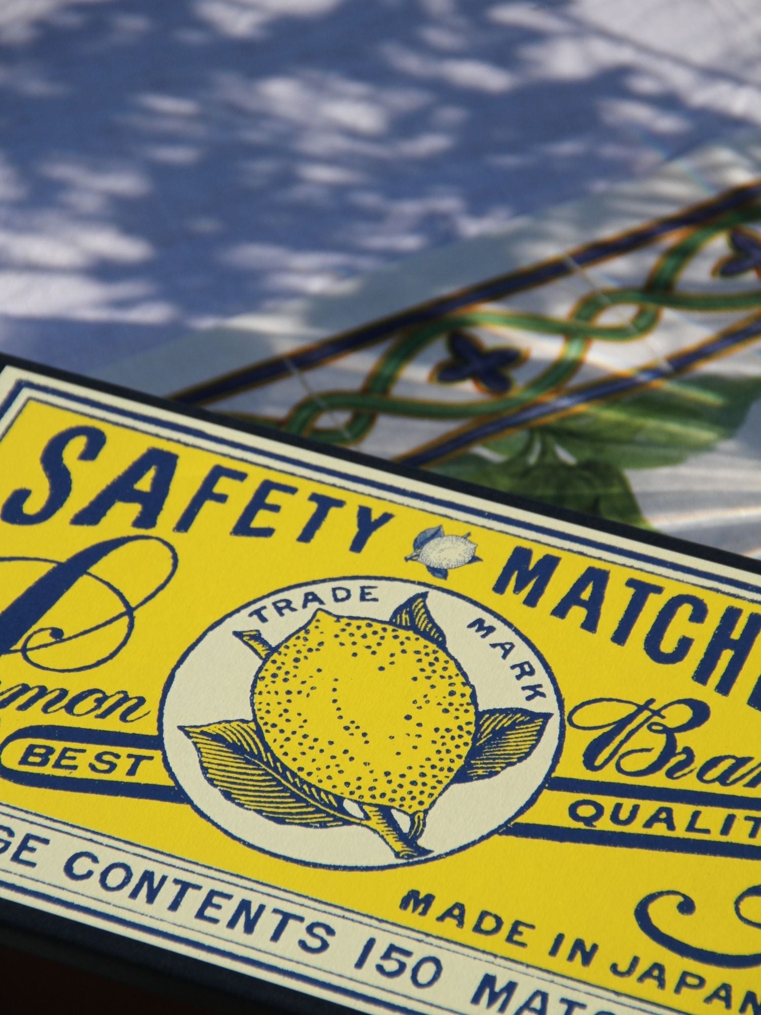 A close-up of a vintage-style yellow safety match box label with blue text and a lemon illustration. The visible text includes 'SAFETY MATCHES', 'Lemon', 'Trade Mark', and 'Made in Japan'.