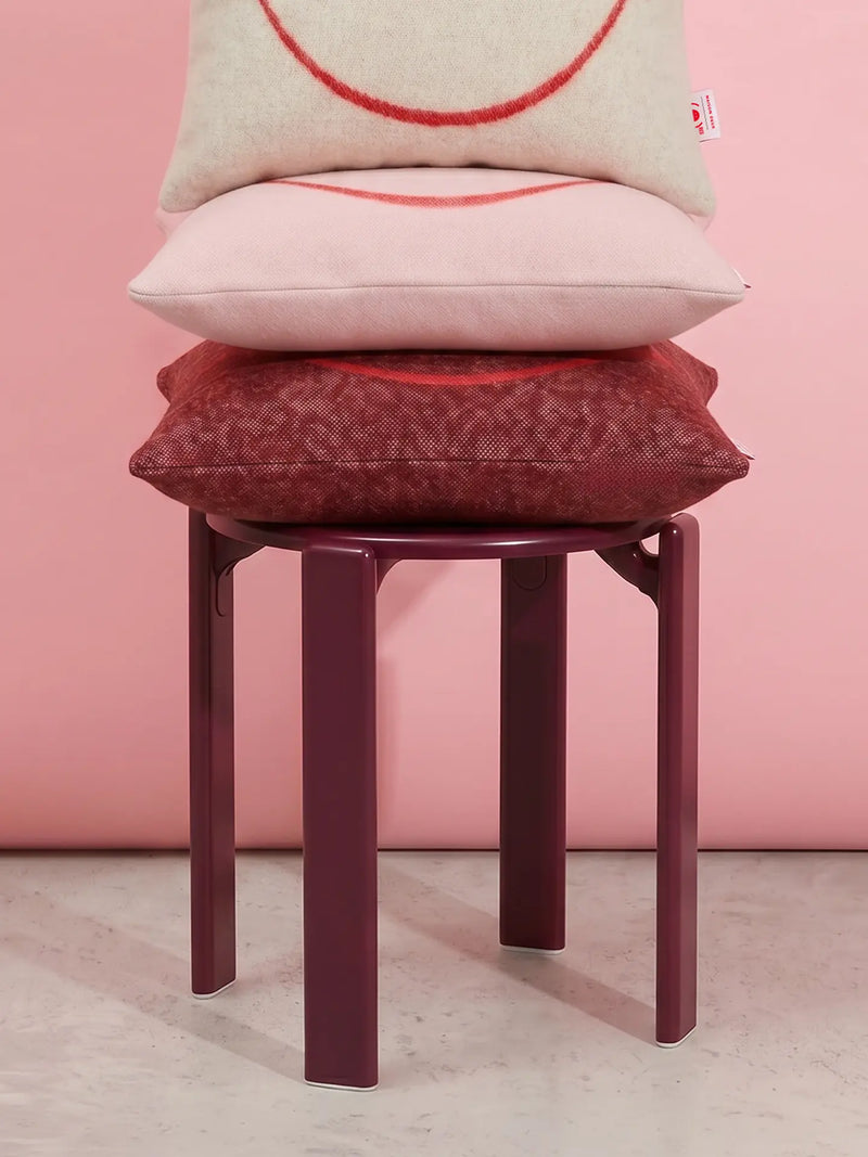 Three decorative throw pillows stacked on a dark burgundy stool against a solid pink wall and light concrete floor. The pillows are cream, pale pink, and deep burgundy with red embroidered smiley faces.