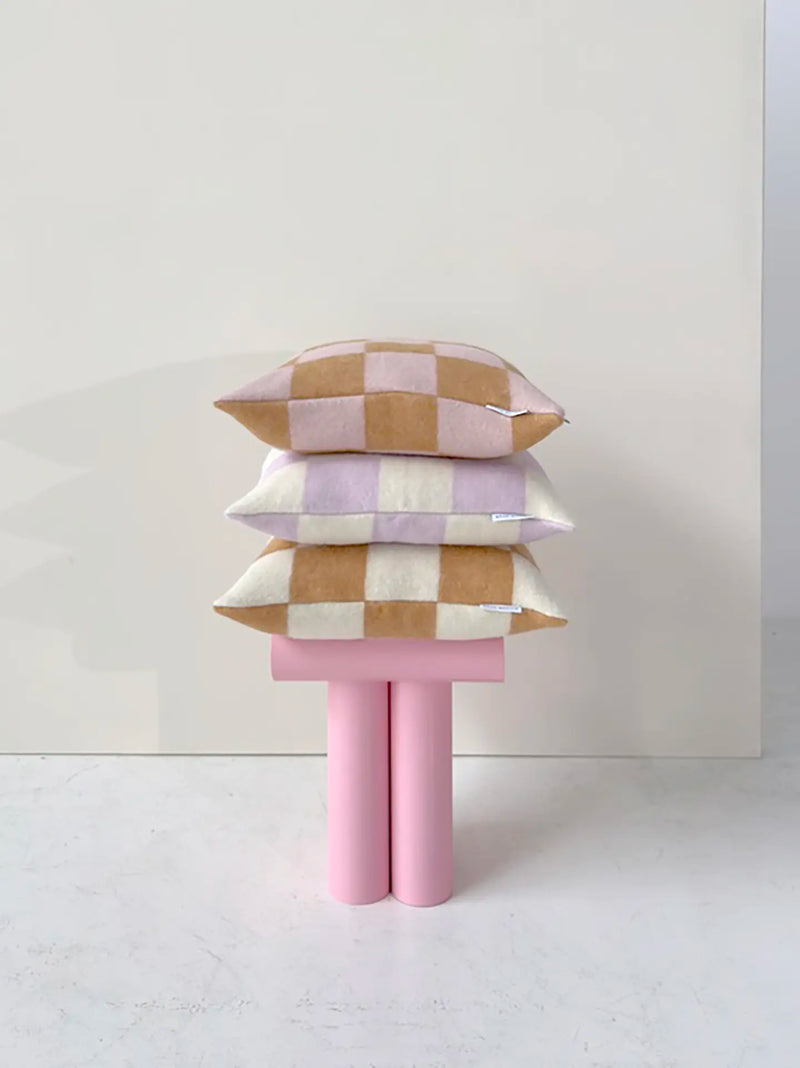 Three checkered throw pillows in beige, pink, and white are stacked on a pink sculptural bench against a minimalist cream wall.