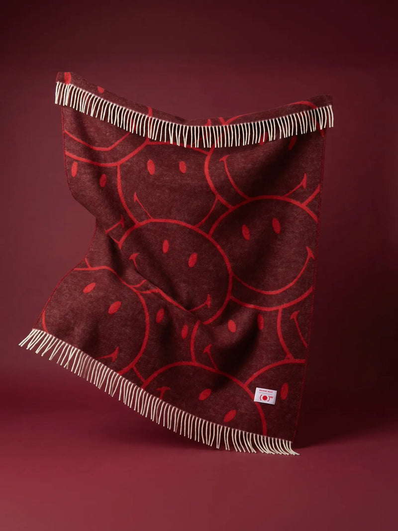 A big dark red woven blanket with a red smiley face pattern and cream-colored fringe, shown draped against a solid dark red background.