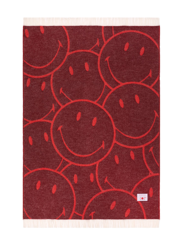 A rectangular woven throw blanket with a red and dark red smiley face pattern, featuring cream-colored fringe on the top and bottom edges and a small white brand tag in the lower right corner.