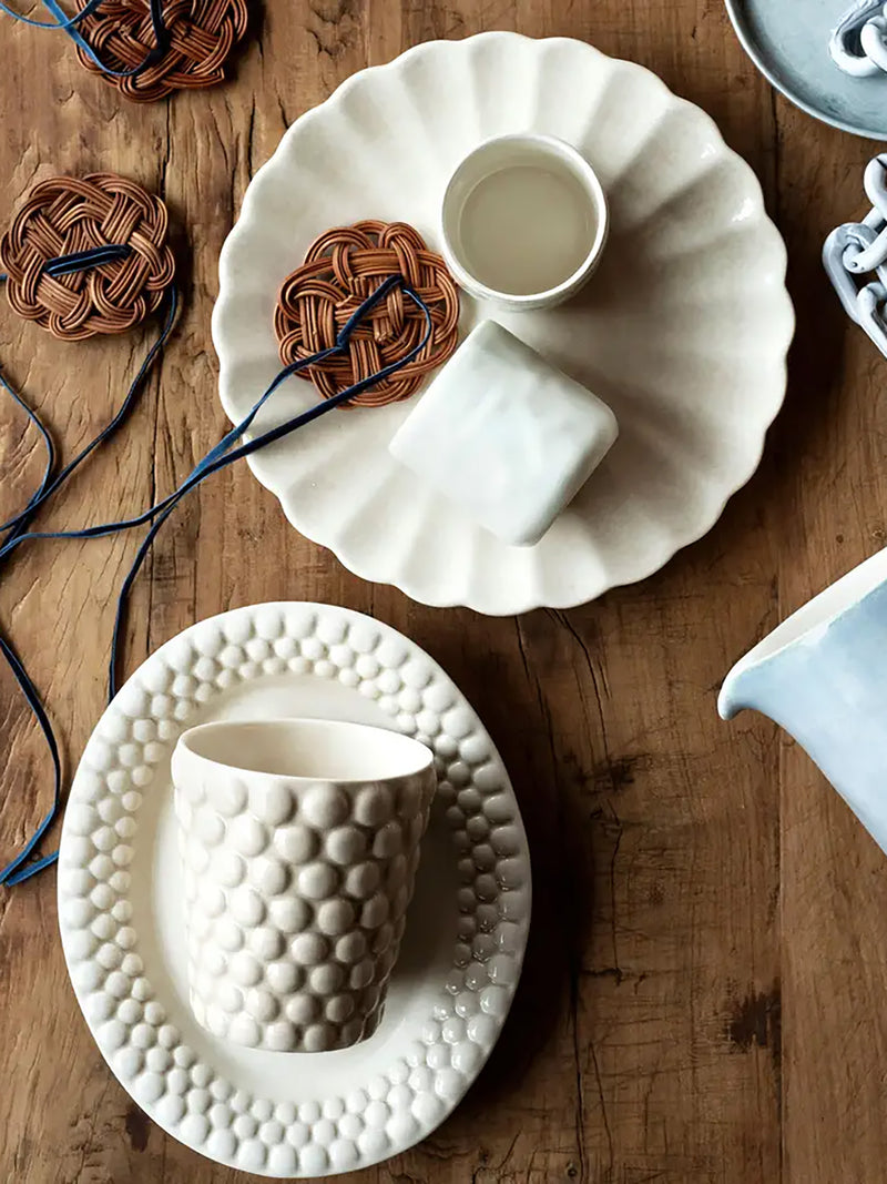Two ceramic cups on textured white plates on a wooden surface with yarn.