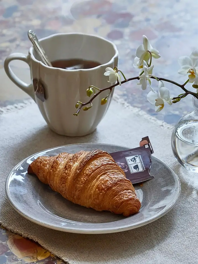 Croissant on a plate with a cup of coffee and a sachet, set against a floral tablecloth.