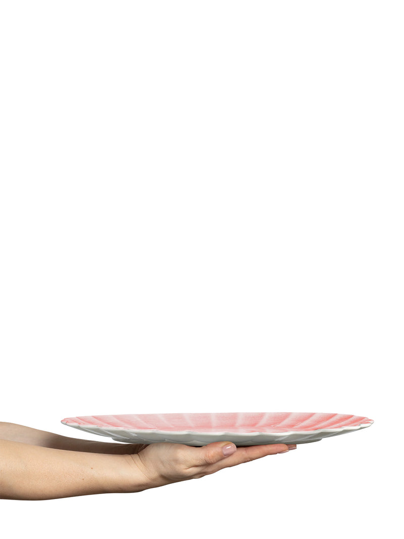 A hand holding a large scalloped ceramic plate with a pink top and white underside against a solid white background.