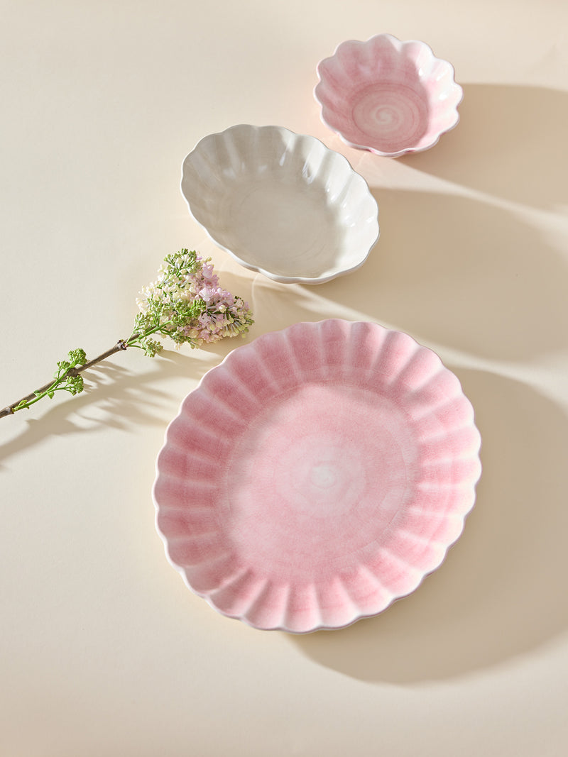 Three scalloped ceramic dishes on a warm cream background: a large pink oval plate in the foreground, a smaller white oval bowl behind it, and a small pink oval bowl at the top right. A delicate flower stem rests beside the dishes.