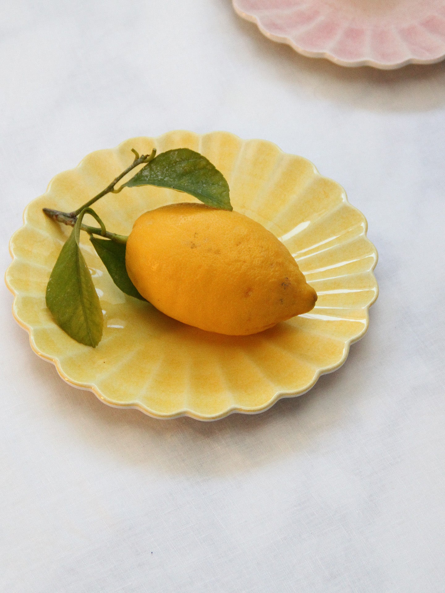 The small Oyster yellow ceramic plate fits  a big lemon, shown here on a light tablecloth with the small Oyster light pink ceramic plate on the backround.