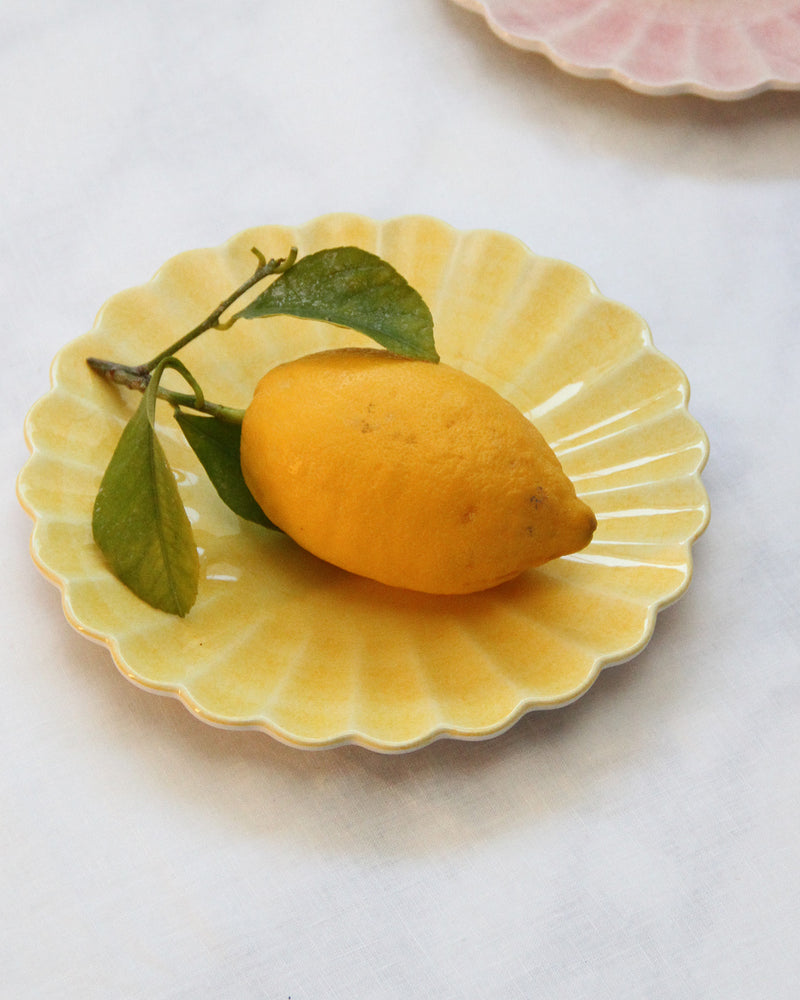 Yellow small ceramic plate from the Oyster collection, with a lemon and green leaf on a white tablecloth and slice of the light pink Oyster plate on the top right corner