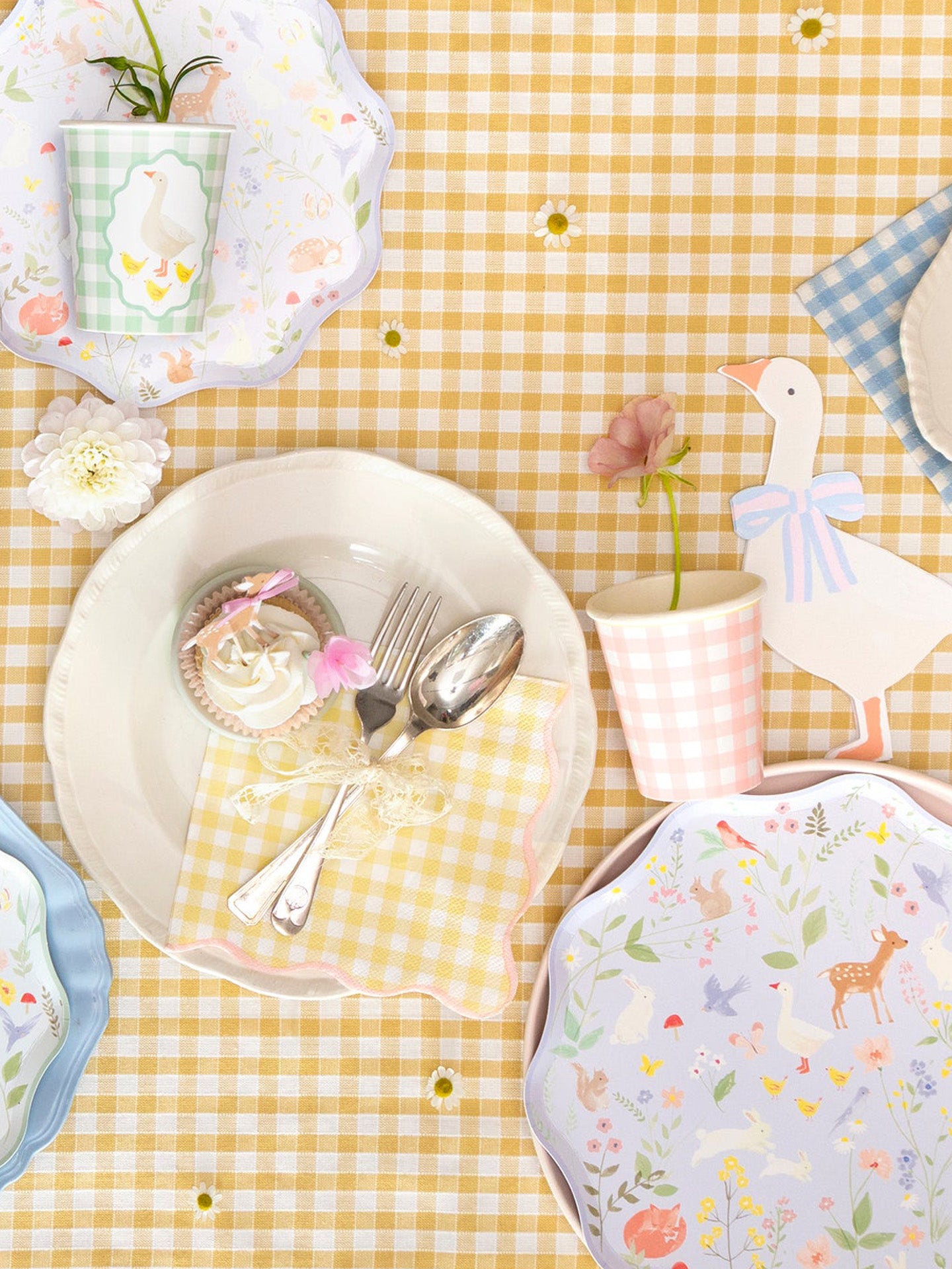 Pastel coloured table setting with decorative plates and gingham patterned cups and dinner napkins, with pretty utensils and cupcakes on a yellow checkered tablecloth.